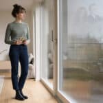 Junge Frau mit alten Familienfotos vor einem Fenster, in dem schemenhaft Menschen aus früheren Generationen zu sehen sind.