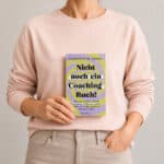 Frau im violetten Pullover hält das Buch „Nicht noch ein Coaching-Buch!“ von Charlotte M. Raven vor neutralem Hintergrund in der Hand.