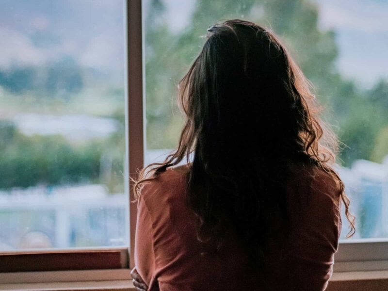 Frau steht mit dem Rücken zur Kamera vor einem Fenster und blickt nach draußen – Symbolbild für Zweifel und stille Erwartungen rund um Manifestation.